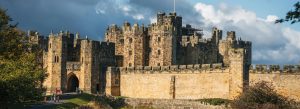 Medieval stone castle with fortified walls and towers under dramatic sky, likely Alnwick Castle in Northumberland England
