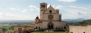 Front view of Basilica of Saint Francis of Assisi overlooking the Umbrian countryside with visitors gathered in the courtyard.