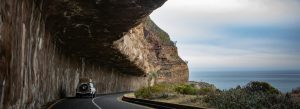 Car driving along Chapman’s Peak Drive under rocky overhang with ocean views near Cape Town