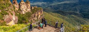 Tourists admiring the iconic Three Sisters rock formation from Echo Point lookout in the Blue Mountains National Park, surrounded by lush forest valleys.