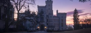 Fairytale-style Hluboká Castle illuminated at dusk with towers and battlements silhouetted against a colorful sunset sky.