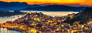Sunset view of a charming coastal town with illuminated houses, marina boats, and calm sea surrounded by mountains
