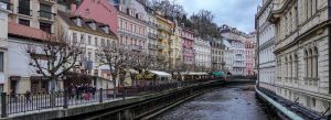 Colorful historic buildings along the Teplá River promenade in Karlovy Vary with cafes and pedestrians strolling beside the water.