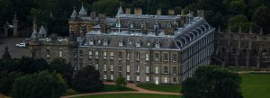 Aerial view of the Palace of Holyroodhouse in Edinburgh, Scotland, surrounded by historic architecture, landscaped grounds, and lush greenery.