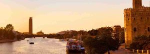 Sunset view of the Guadalquivir River with boats, the historic Torre del Oro on the right, and the modern Torre Sevilla in the background