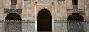 Decorative courtyard entrance inside Al-Attarine Madrasa in Fes, Morocco, featuring carved cedar wood, stucco, and colorful zellij tiles.