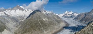 Aerial panorama of the Aletsch Glacier flowing through snow-covered Swiss Alps under a bright blue sky.