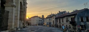 Sunset view beside the ancient Roman Arles Amphitheatre in Arles, France, with historic stone arches and traditional French buildings lining the street.