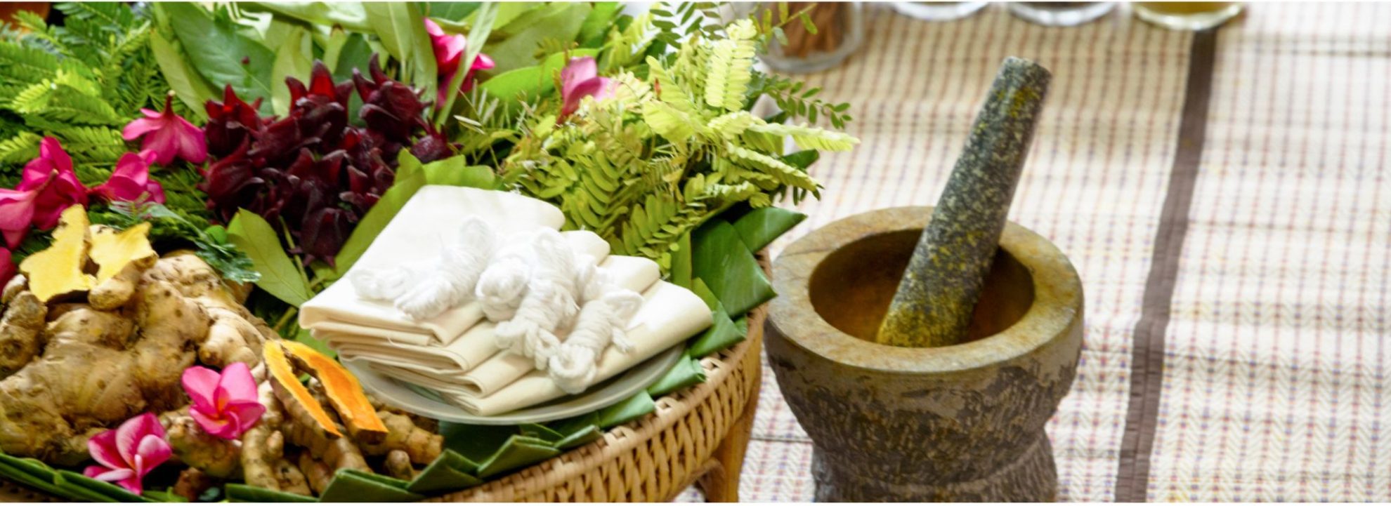 Ayursoma traditional herbal wellness setup with tropical flowers, herbal compress balls, ginger, turmeric, and stone mortar for authentic Ayurvedic spa experiences.