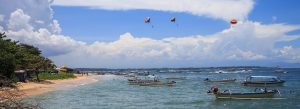 Traditional boats and parasailing activities at Benoa Beach in Bali, Indonesia, under a bright blue sky with calm tropical waters.