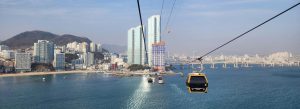 Aerial view of Songdo Marine Cable Car gliding over the ocean toward Busan skyline with high-rise buildings, beach, and coastal bridge.