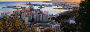 Panoramic view of Málaga, Spain featuring the Mediterranean coastline, harbor, city skyline, and historic bullring at sunset.