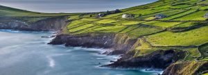 Scenic coastal view of the Dingle Peninsula in Ireland with green patchwork fields, rugged cliffs, stone walls, and Atlantic Ocean waves at sunset.