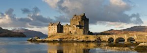 Historic Eilean Donan Castle standing beside a stone bridge with mountains and calm water in the Scottish Highlands.