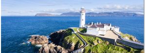 Fanad Head Lighthouse on the rocky coastline of County Donegal, Ireland, overlooking the Atlantic Ocean under a bright blue sky.