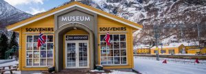 Exterior of the Flåm Railway Museum in Norway with snow-covered mountains, Norwegian flags, and railway tracks nearby.