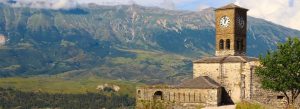 Historic stone church and clock tower in Gjirokastra, Albania, with dramatic mountain scenery in the background.