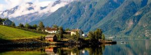 Panoramic view of Hardangerfjord in Norway with peaceful waterfront cottages, green hills, calm reflective waters, and snow-capped mountains under drifting clouds.
