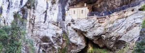 The Holy Cave of Covadonga built into a dramatic rocky cliff in Asturias, Spain, with visitors walking along a scenic mountain pathway.
