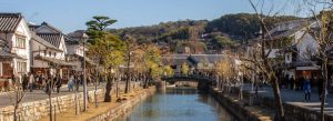 Traditional canal-lined street in Kurashiki Bikan Historical Quarter, Okayama Prefecture, Japan, with white-walled Edo-period buildings and stone bridge.