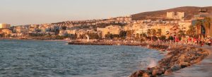 Sunset view of Kusadasi coastal town in Turkey with seaside promenade, rocky shoreline, and hillside cityscape overlooking the Aegean Sea.