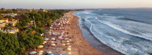 Aerial view of Kuta Beach in Bali with golden sand, colorful beach umbrellas, ocean waves, and tourists enjoying the tropical coastline at sunset.