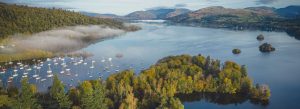Aerial view of Lake Windermere surrounded by forests, sailboats, and rolling hills in England’s Lake District.