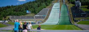 Family overlooking the Lysgårdsbakkene Ski Jumping Arena in Lillehammer, Norway with towering Olympic ski jumps surrounded by forested hills.