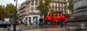 Street view of Montmartre in Paris, France, featuring classic Parisian buildings, cafés, cyclists, and wet streets on a cloudy day.