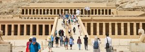 Tourists walking toward the Mortuary Temple of Hatshepsut in Luxor, Egypt, surrounded by dramatic desert cliffs and ancient colonnades.