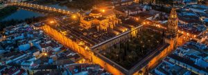 Aerial evening view of the Mosque-Cathedral of Córdoba in Spain illuminated with golden lights beside the Guadalquivir River and Roman Bridge.