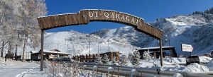Snow-covered entrance arch of Oi-Qaragai Mountain Resort in Kazakhstan with ski slopes and mountains in the background.