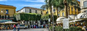 Piazza Tasso in Sorrento, Italy with palm trees, cafés, tourists, and colorful Mediterranean buildings on a sunny day.