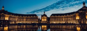 Illuminated historic buildings at Place de la Bourse in Bordeaux, France, reflected in the water at dusk under a dramatic evening sky.