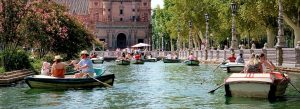 Visitors rowing boats along the canal at Plaza de España in Seville, Spain, surrounded by ornate bridges, ceramic details, and lush trees on a sunny day.