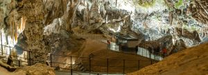 Interior view of Postojna Cave in Slovenia featuring massive stalactites, stalagmites, and illuminated underground pathways.
