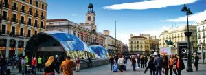 Puerta del Sol square in Madrid, Spain, featuring the modern glass metro entrance and historic architecture with tourists walking through the city center.