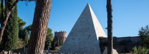Ancient Pyramid of Cestius in Rome, Italy, surrounded by tall pine trees and historic Roman architecture under a clear blue sky.
