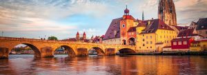 Historic stone bridge and colorful medieval buildings along the Danube River in Regensburg, Germany at sunset.