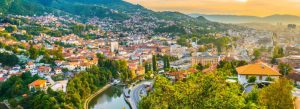 Panoramic view of Sarajevo, Bosnia and Herzegovina, with rolling hills, river scenery, and colorful rooftops at sunset.