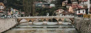 Historic stone bridge over the Miljacka River in Sarajevo, Bosnia and Herzegovina, surrounded by traditional houses and hillside scenery.