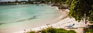 Tranquil sandy beach in Barbados with clear turquoise water, a lounge chair under leafy trees, and small boats anchored near the shore