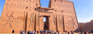 Front entrance of the ancient Temple of Edfu in Egypt featuring massive sandstone walls covered with hieroglyphics and visitors exploring the site.