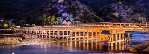 Illuminated Togetsukyo Bridge crossing the Katsura River in Arashiyama, Kyoto, with colorful spring trees glowing at night.