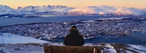 Snow-covered panoramic view of Tromsø, Norway with Arctic mountains, fjords, and a traveler overlooking the city from a scenic viewpoint