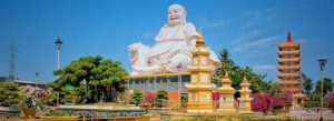 Vinh Trang Pagoda in My Tho, Vietnam featuring the giant white Laughing Buddha statue, colorful gardens, and traditional pagoda tower under a blue sky.