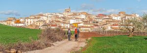 Hikers walking along a rural trail toward a traditional village in Galicia, Spain, during a Camino pilgrimage route.