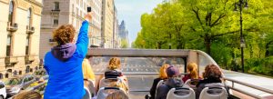 Tourists riding an open-top sightseeing bus along Central Park in New York City on a sunny day.